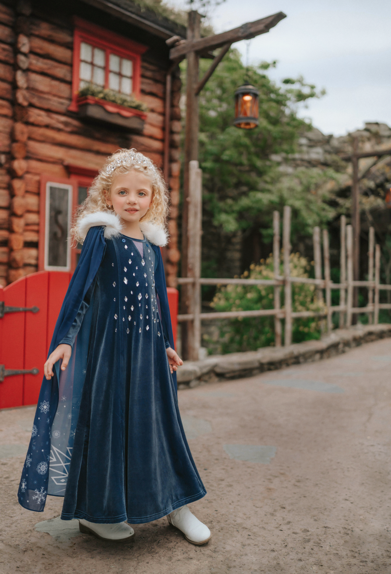 A young girl is wearing a navy blue velvet gown is adorned with diamond shape crystal gems of various sizes.  There is a small lavender triangle near the neck. She has on a navy blue chiffon cape with white faux fur trim on the collar. There is a snow flake design on the back of the cape.