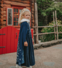 A young girl is wearing a navy blue velvet gown is adorned with diamond shape crystal gems of various sizes.  There is a small lavender triangle near the neck. She has on a navy blue chiffon cape with white faux fur trim on the collar. There is a snow flake design on the back of the cape.