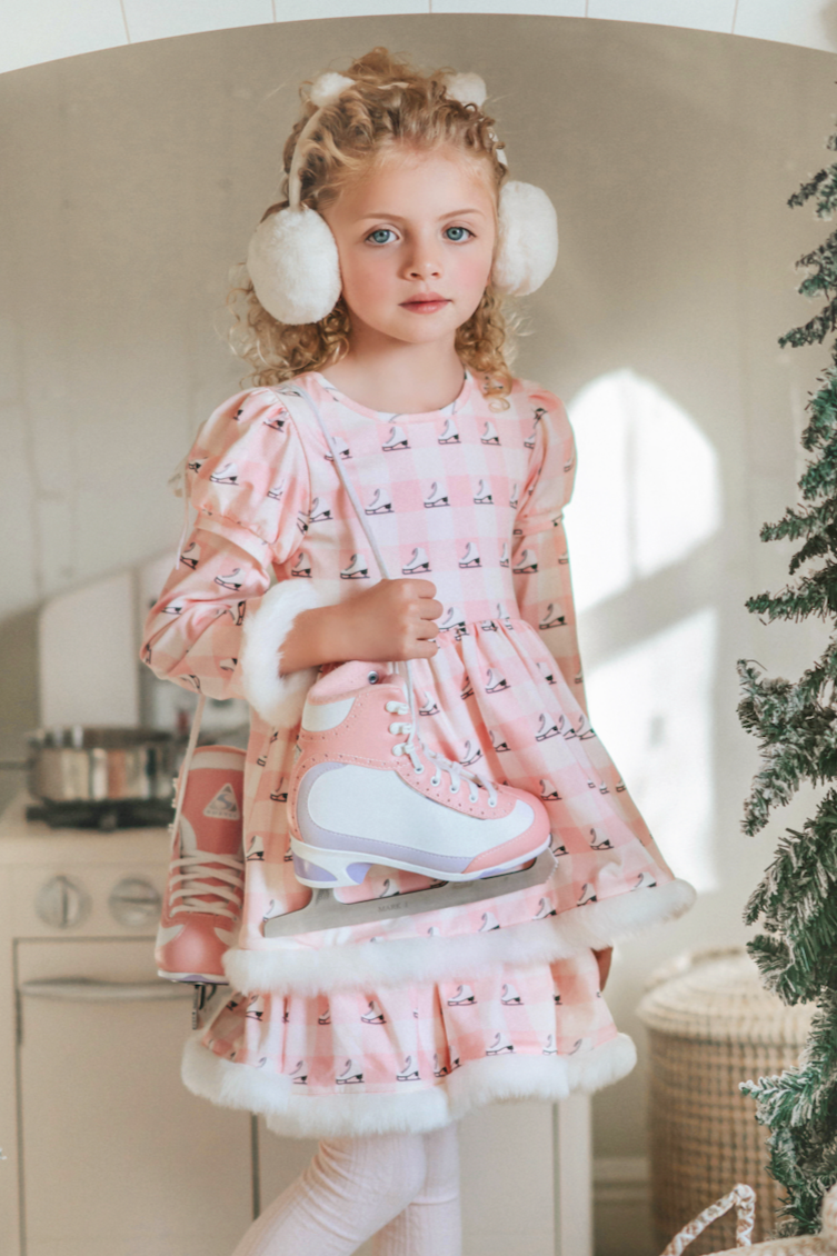 A young girl is wearing a long sleeve pink twirl dress adorned with white and silver ice skates.  The sleeves and hem of the twirl are lined with white faux fur.  There is a double layer skirt for excellent twirling.  Also included is a white faux fur hand muff with pink bow. 