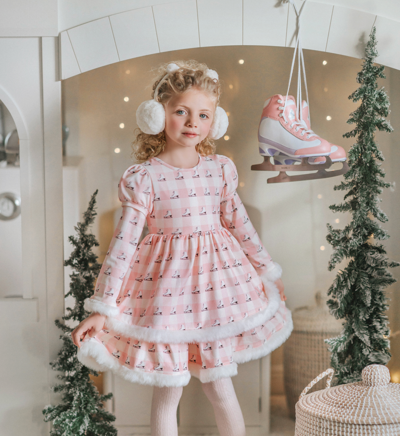 A young girl is wearing a long sleeve pink twirl dress adorned with white and silver ice skates.  The sleeves and hem of the twirl are lined with white faux fur.  There is a double layer skirt for excellent twirling.  Also included is a white faux fur hand muff with pink bow. 