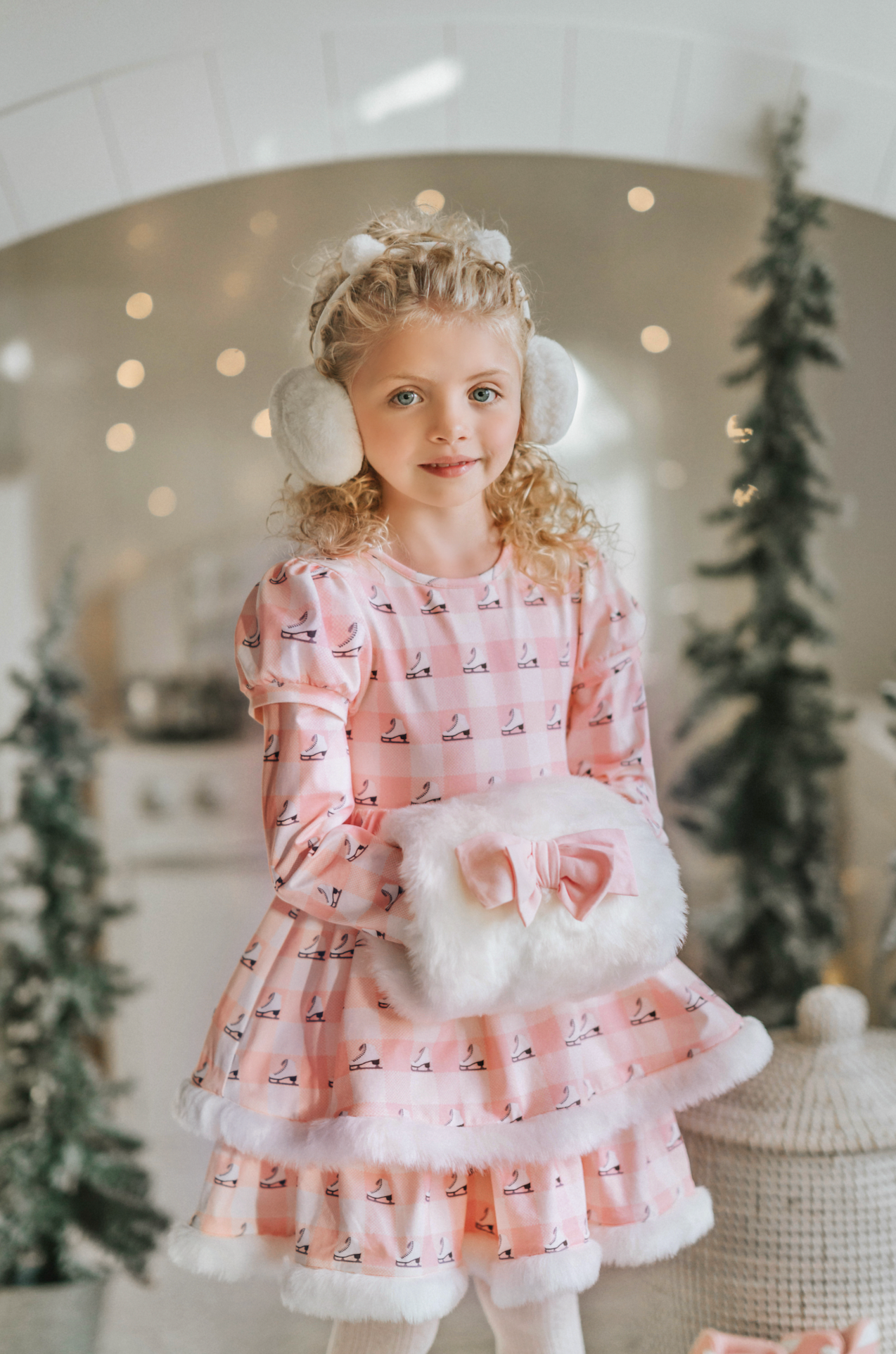 A young girl is wearing a long sleeve pink twirl dress adorned with white and silver ice skates.  The sleeves and hem of the twirl are lined with white faux fur.  There is a double layer skirt for excellent twirling.  Also included is a white faux fur hand muff with pink bow. 