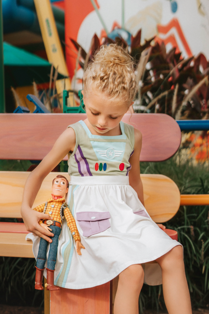 A young girl wears a sporty space hero twirl dress.  The cotton dress is sleeveless and has light green panels on the bodice with embroidered button like features.  There are light green and light purple racing stripes down the side. The front has two pockets on the skirt. 