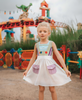 A young girl wears a sporty space hero twirl dress.  The cotton dress is sleeveless and has light green panels on the bodice with embroidered button like features.  There are light green and light purple racing stripes down the side. The front has two pockets on the skirt. 