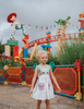 A young girl wears a sporty space hero twirl dress.  The cotton dress is sleeveless and has light green panels on the bodice with embroidered button like features.  There are light green and light purple racing stripes down the side. The front has two pockets on the skirt. 