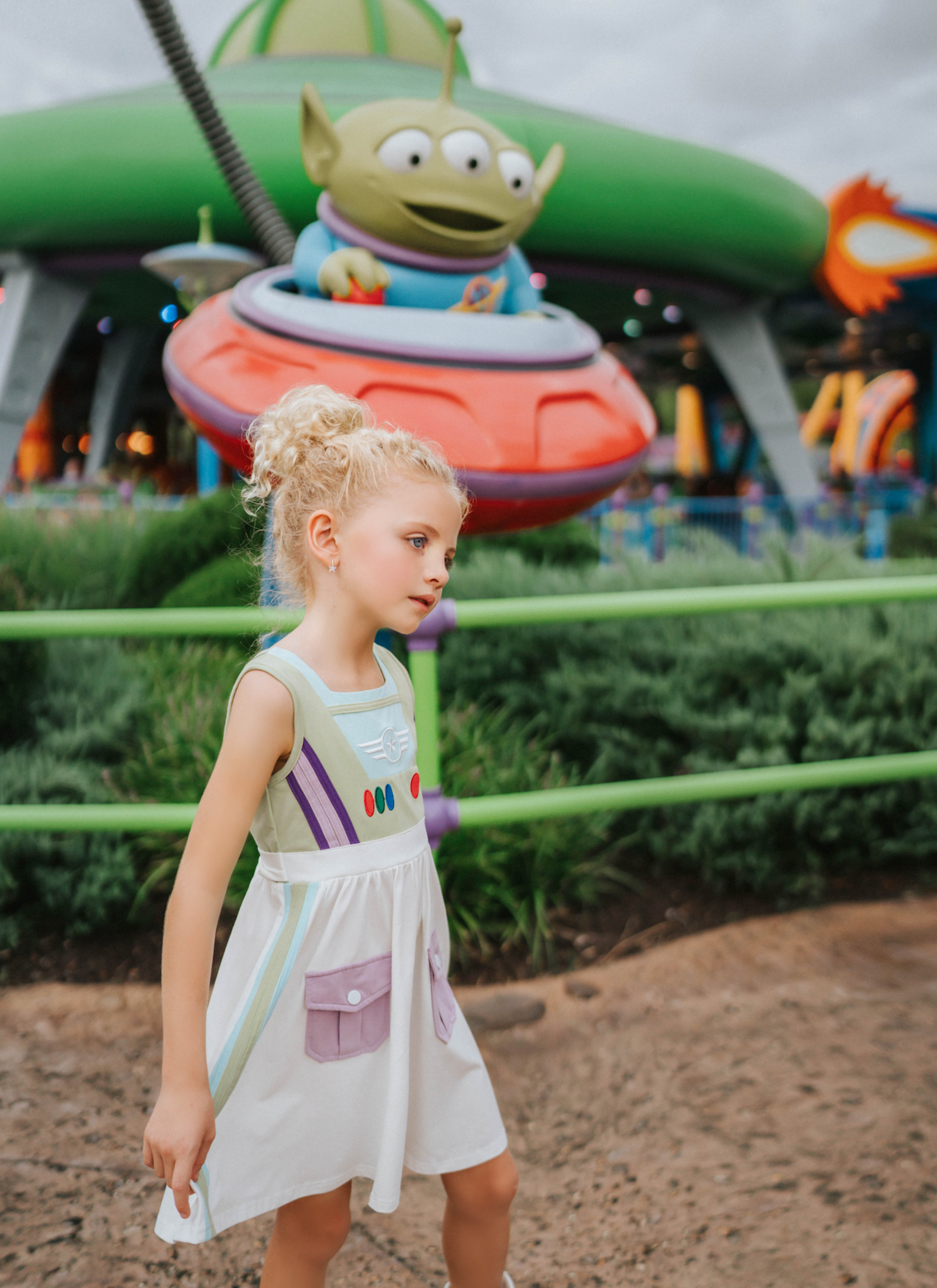 A young girl wears a sporty space hero twirl dress.  The cotton dress is sleeveless and has light green panels on the bodice with embroidered button like features.  There are light green and light purple racing stripes down the side. The front has two pockets on the skirt. 