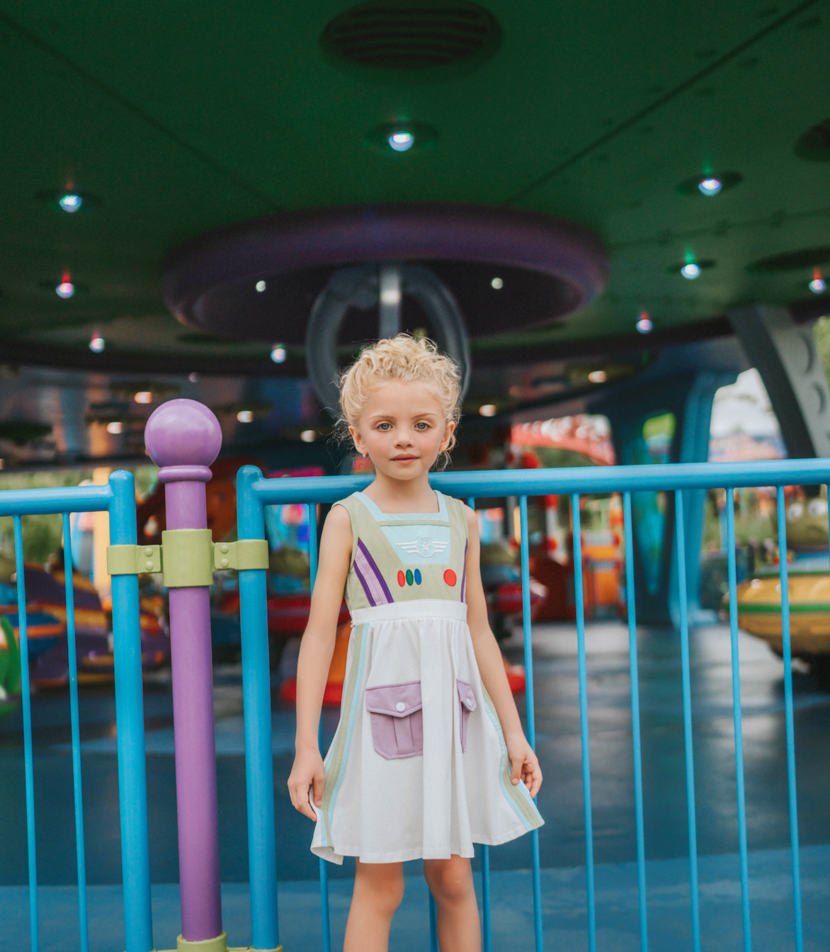 A young girl wears a sporty space hero twirl dress.  The cotton dress is sleeveless and has light green panels on the bodice with embroidered button like features.  There are light green and light purple racing stripes down the side. The front has two pockets on the skirt. 