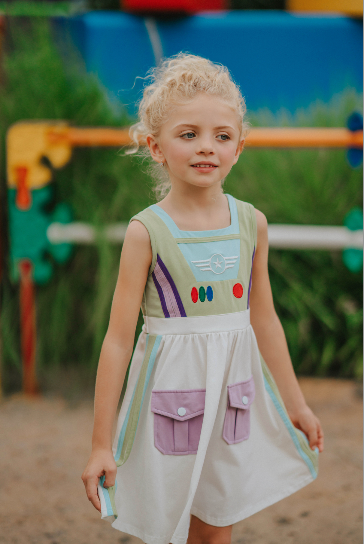 A young girl wears a sporty space hero twirl dress.  The cotton dress is sleeveless and has light green panels on the bodice with embroidered button like features.  There are light green and light purple racing stripes down the side. The front has two pockets on the skirt. 