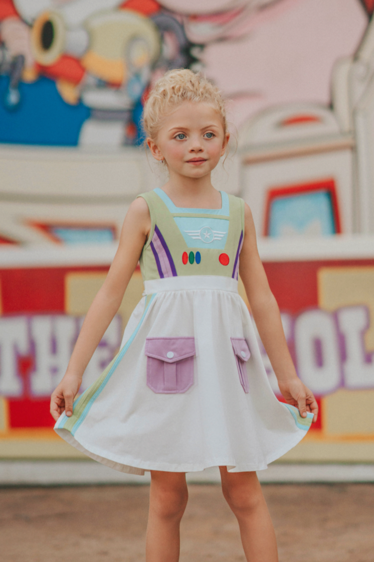 A young girl wears a sporty space hero twirl dress.  The cotton dress is sleeveless and has light green panels on the bodice with embroidered button like features.  There are light green and light purple racing stripes down the side. The front has two pockets on the skirt. 