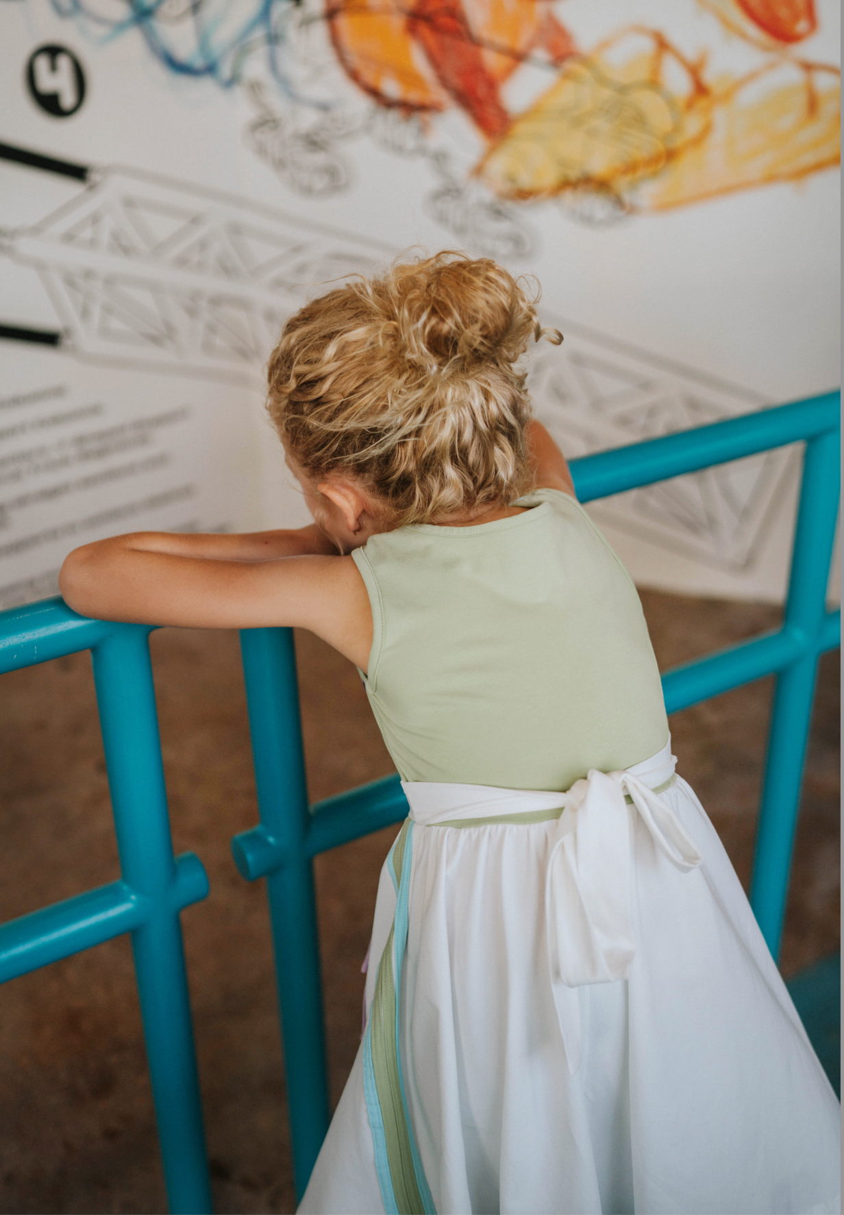 A young girl wears a sporty space hero twirl dress.  The cotton dress is sleeveless and has light green panels on the bodice with embroidered button like features.  There are light green and light purple racing stripes down the side. The front has two pockets on the skirt. 