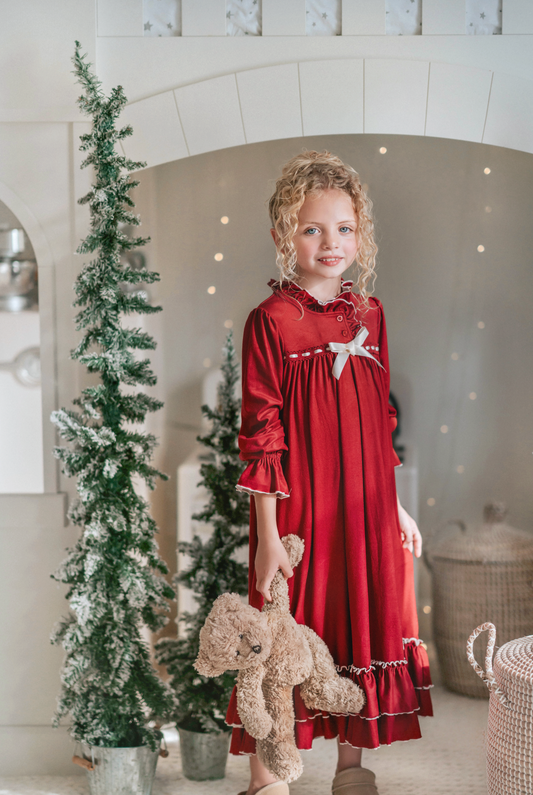 A young girl is wearing The Original Clara Nutcracker Gown in Merry Maroon, featuring an ivory bow. The buttery soft fabric is cozy and sensory friendly.  The long sleeves and the ruffles at the hem add a traditional classic look.