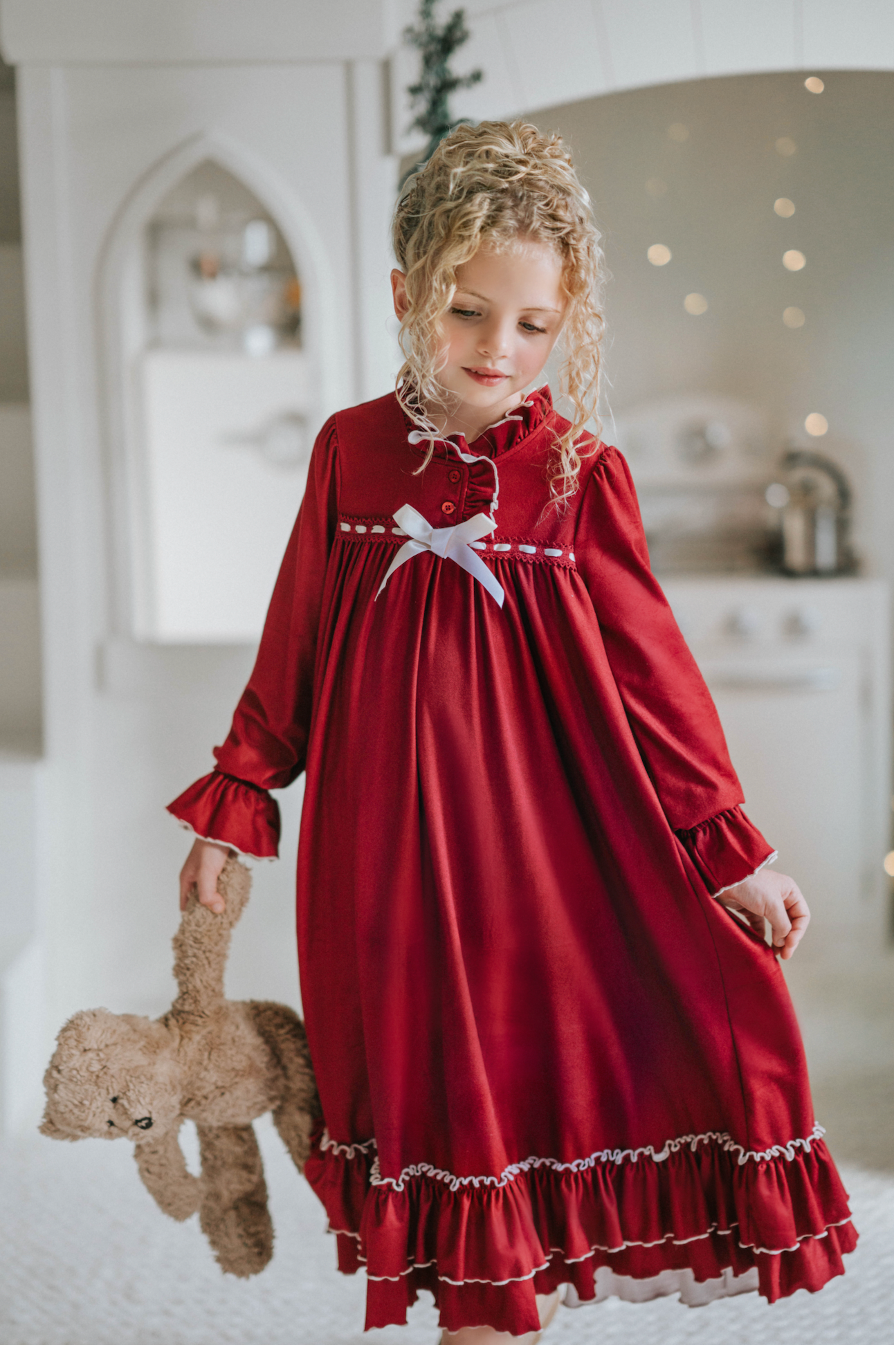 A young girl is wearing The Original Clara Nutcracker Gown in Merry Maroon, featuring an ivory bow. The buttery soft fabric is cozy and sensory friendly.  The long sleeves and the ruffles at the hem add a traditional classic look.