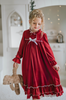 A young girl is wearing The Original Clara Nutcracker Gown in Merry Maroon, featuring an ivory bow. The buttery soft fabric is cozy and sensory friendly.  The long sleeves and the ruffles at the hem add a traditional classic look.