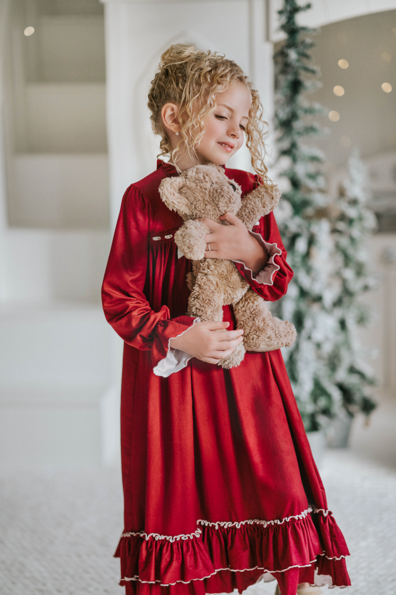 A young girl is wearing The Original Clara Nutcracker Gown in Merry Maroon, featuring an ivory bow. The buttery soft fabric is cozy and sensory friendly.  The long sleeves and the ruffles at the hem add a traditional classic look.