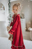 A young girl is wearing The Original Clara Nutcracker Gown in Merry Maroon, featuring an ivory bow. The buttery soft fabric is cozy and sensory friendly.  The long sleeves and the ruffles at the hem add a traditional classic look.