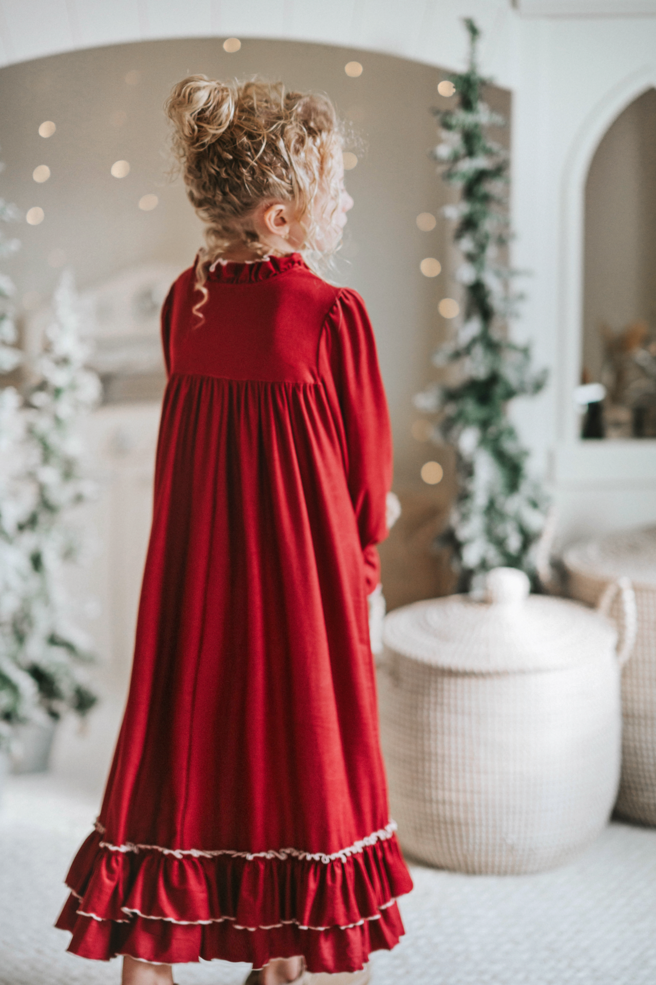 A young girl is wearing The Original Clara Nutcracker Gown in Merry Maroon, featuring an ivory bow. The buttery soft fabric is cozy and sensory friendly.  The long sleeves and the ruffles at the hem add a traditional classic look.