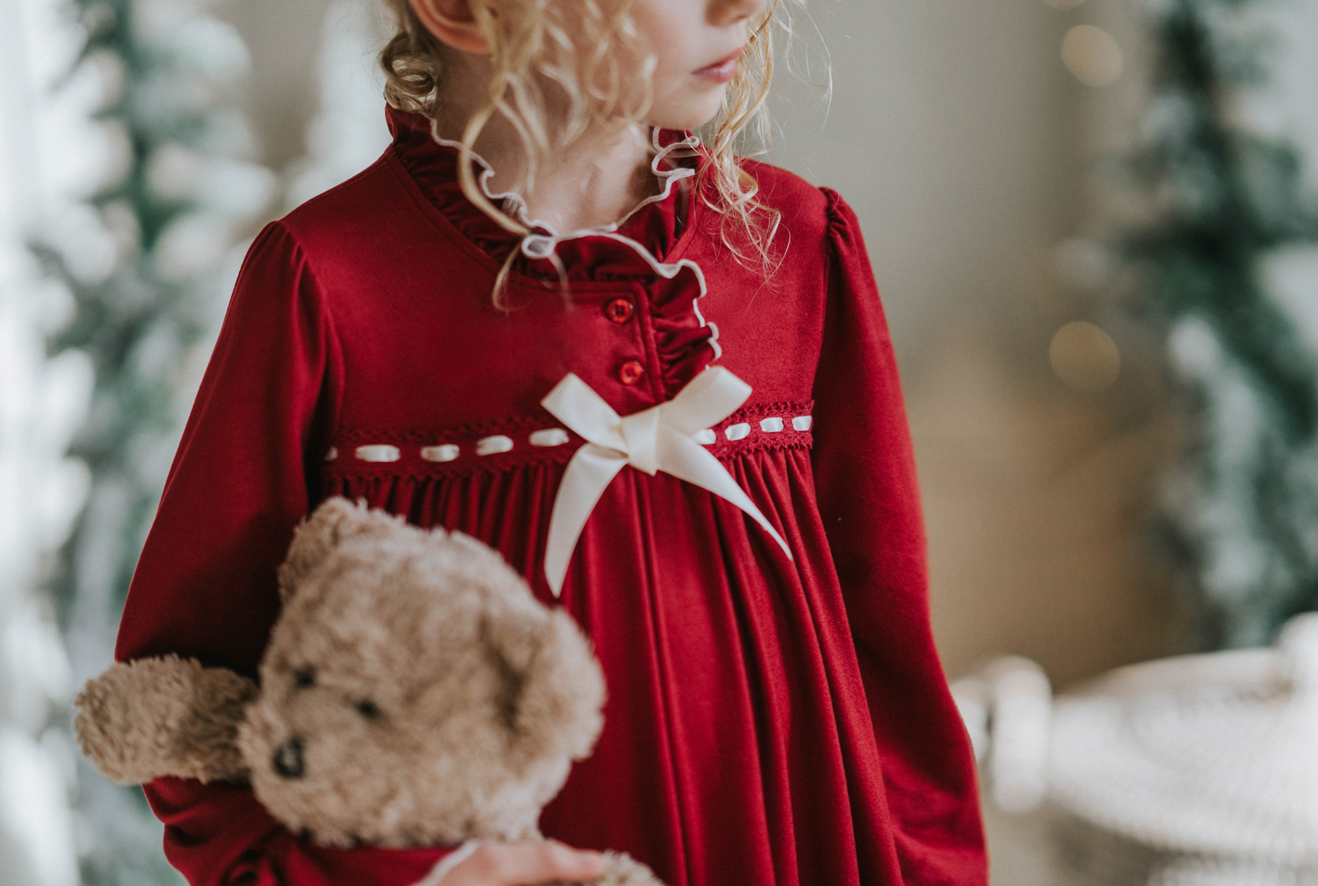 A young girl is wearing The Original Clara Nutcracker Gown in Merry Maroon, featuring an ivory bow. The buttery soft fabric is cozy and sensory friendly.  The long sleeves and the ruffles at the hem add a traditional classic look.