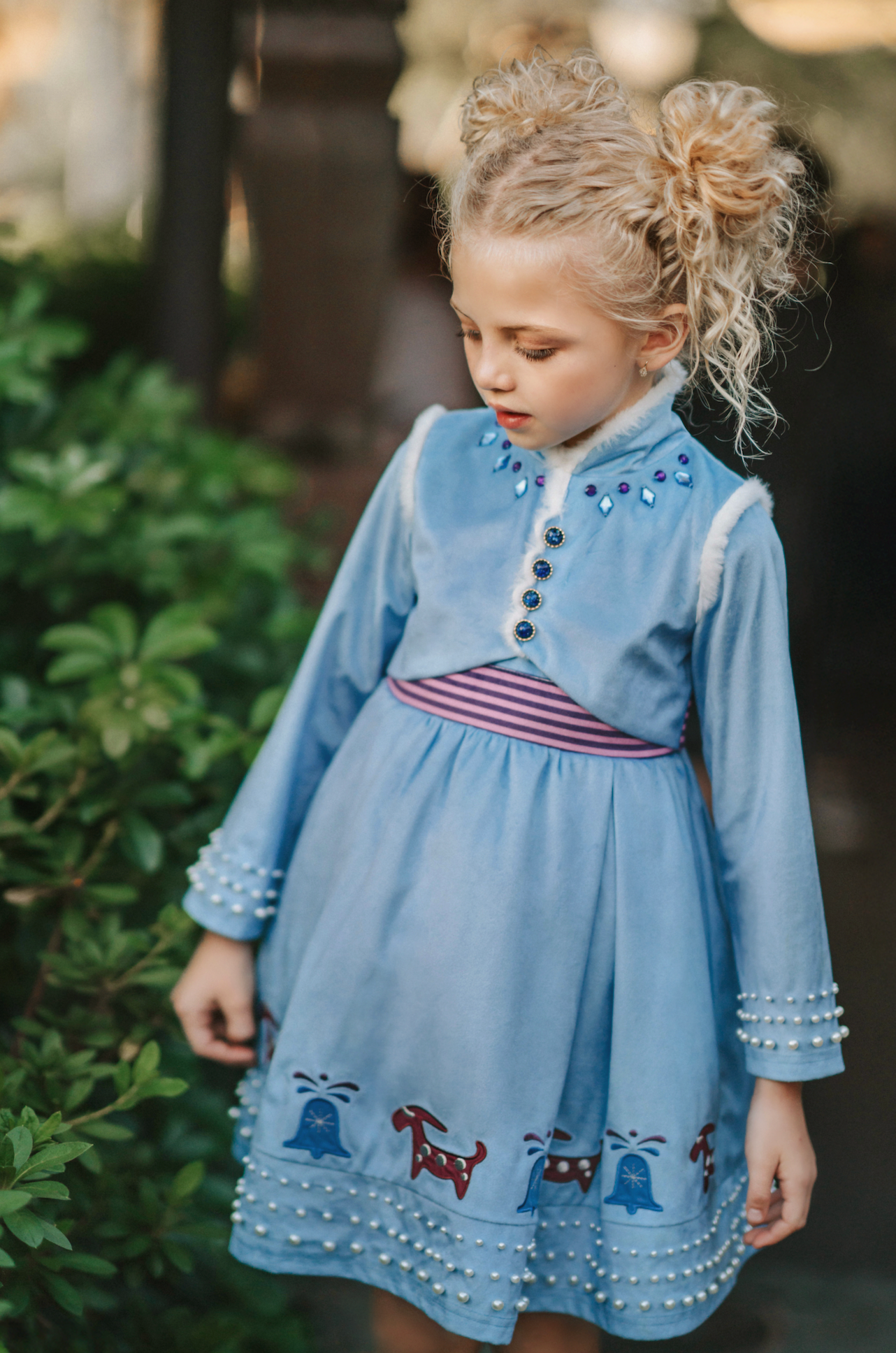 A young girl is wearing a beautiful blue velvet dress.  There is white faux fur trim on the collar and attached jacket. Adorned with blue and purple crystal gems and intricate buttons. The bottom of the dress has a blue bell and Scandinavian mountain goat applique. There are three rows of pearls on the sleeves and the hem of the skirts. 