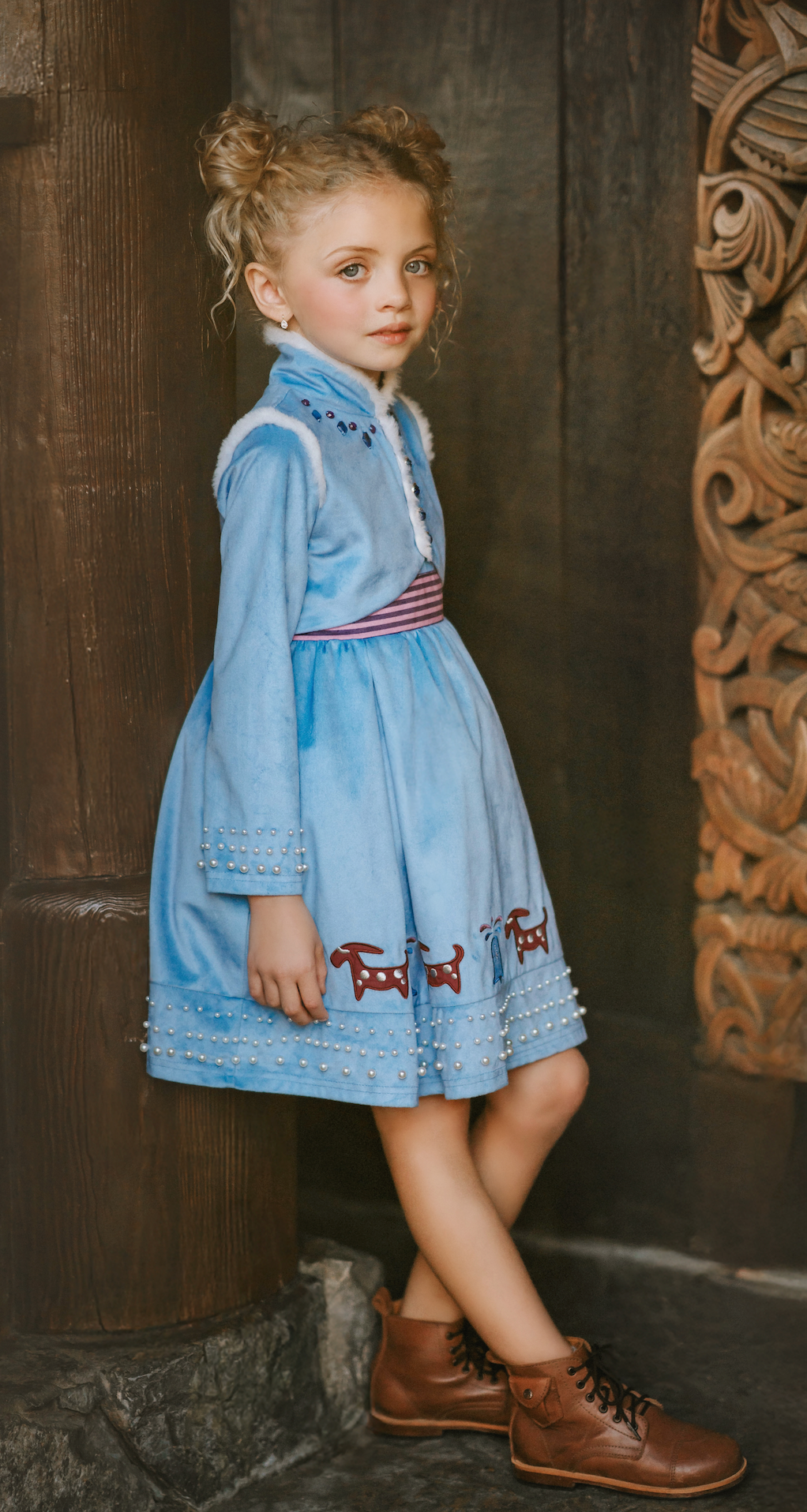 A young girl is wearing a beautiful blue velvet dress.  There is white faux fur trim on the collar and attached jacket. Adorned with blue and purple crystal gems and intricate buttons. The bottom of the dress has a blue bell and Scandinavian mountain goat applique. There are three rows of pearls on the sleeves and the hem of the skirts. 