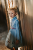 A young girl is wearing a beautiful blue velvet dress.  There is white faux fur trim on the collar and attached jacket. Adorned with blue and purple crystal gems and intricate buttons. The bottom of the dress has a blue bell and Scandinavian mountain goat applique. There are three rows of pearls on the sleeves and the hem of the skirts. 