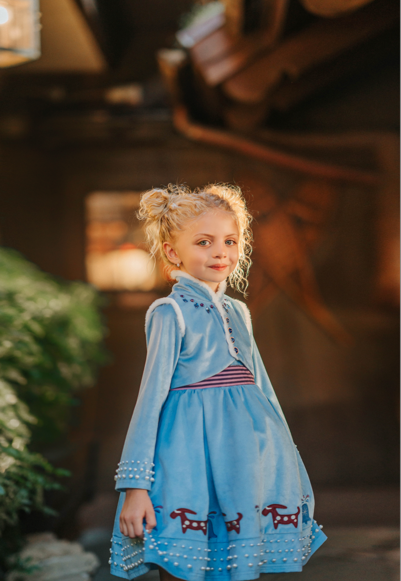 A young girl is wearing a beautiful blue velvet dress.  There is white faux fur trim on the collar and attached jacket. Adorned with blue and purple crystal gems and intricate buttons. The bottom of the dress has a blue bell and Scandinavian mountain goat applique. There are three rows of pearls on the sleeves and the hem of the skirts. 