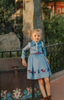 A young girl is wearing a beautiful blue velvet dress.  There is white faux fur trim on the collar and attached jacket. Adorned with blue and purple crystal gems and intricate buttons. The bottom of the dress has a blue bell and Scandinavian mountain goat applique. There are three rows of pearls on the sleeves and the hem of the skirts. 