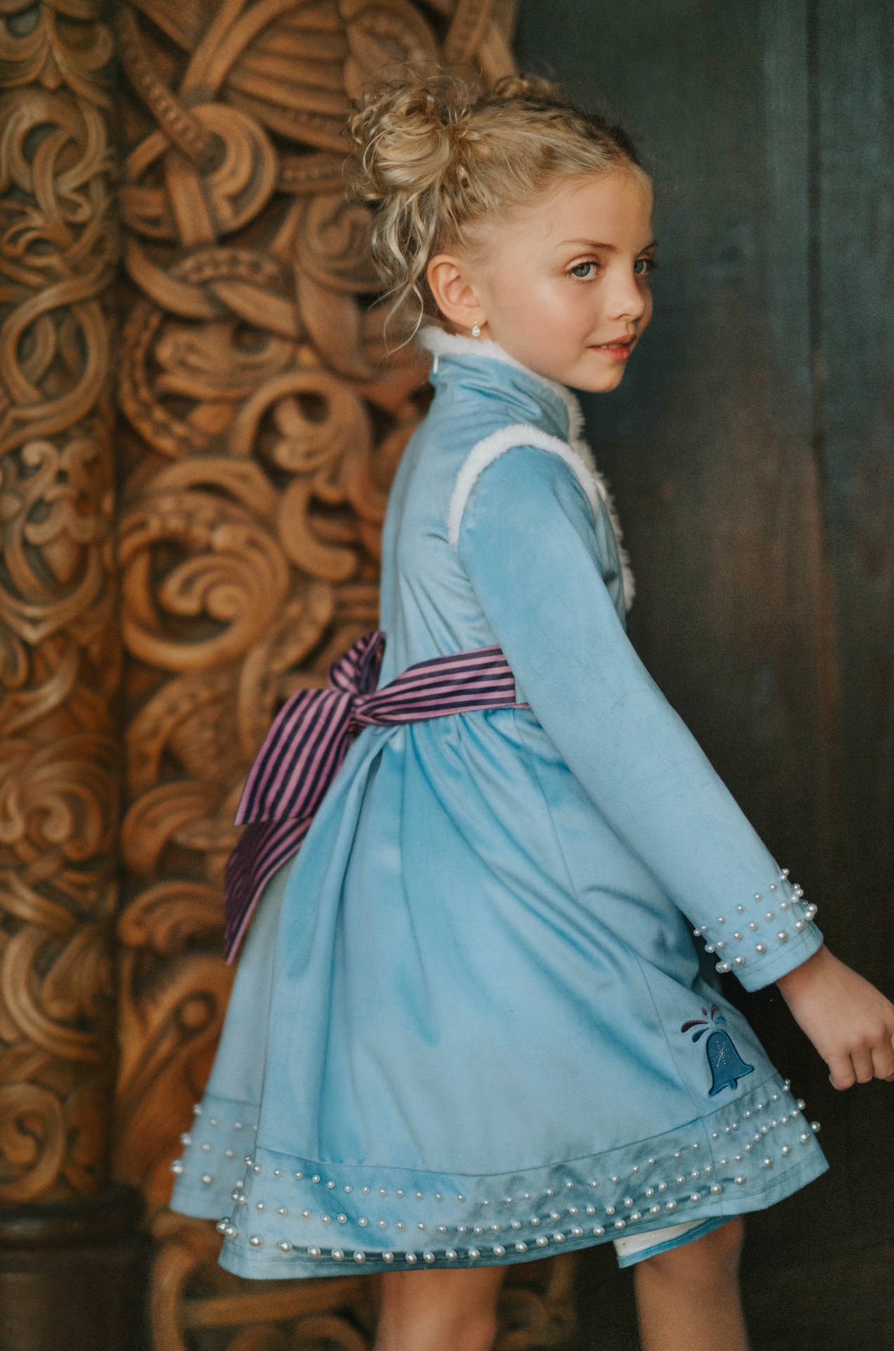 A young girl is wearing a beautiful blue velvet dress.  There is white faux fur trim on the collar and attached jacket. Adorned with blue and purple crystal gems and intricate buttons. The bottom of the dress has a blue bell and Scandinavian mountain goat applique. There are three rows of pearls on the sleeves and the hem of the skirts. 