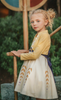 A young girl stands wearing a cotton twirl dress. There is sewn in petti skirt for volume. A geometric golden detail adorns the ivory dress.  There is a purple band with tie at the waist.  The sleevless dress comes with a golden yellow bolero with print on the front in true Arendale princess fashion.