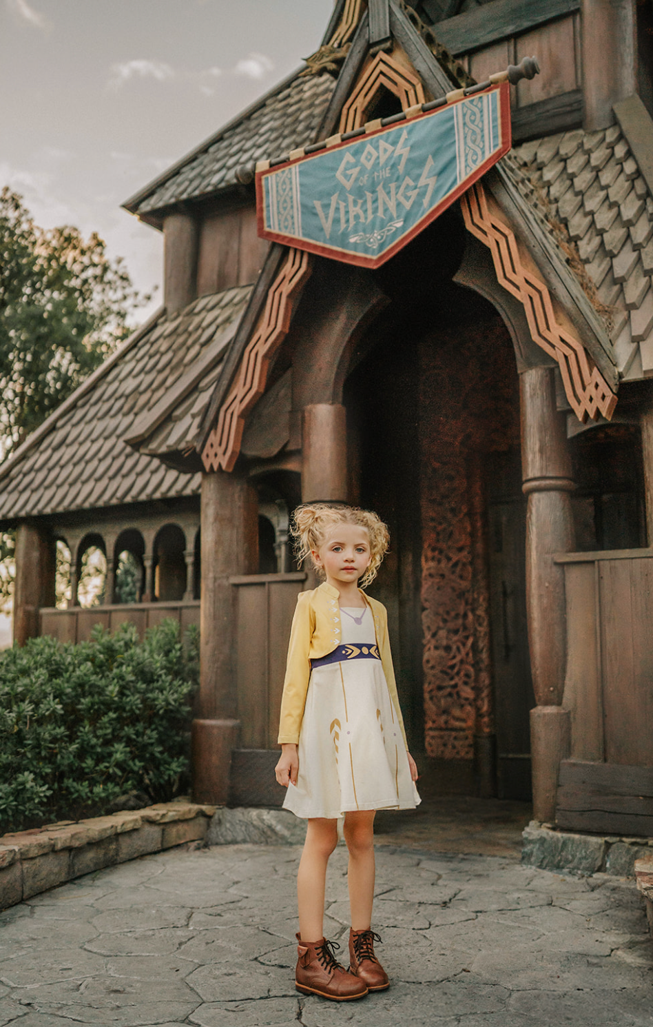 A young girl stands wearing a cotton twirl dress. There is sewn in petti skirt for volume. A geometric golden detail adorns the ivory dress.  There is a purple band with tie at the waist.  The sleevless dress comes with a golden yellow bolero with print on the front in true Arendale princess fashion.