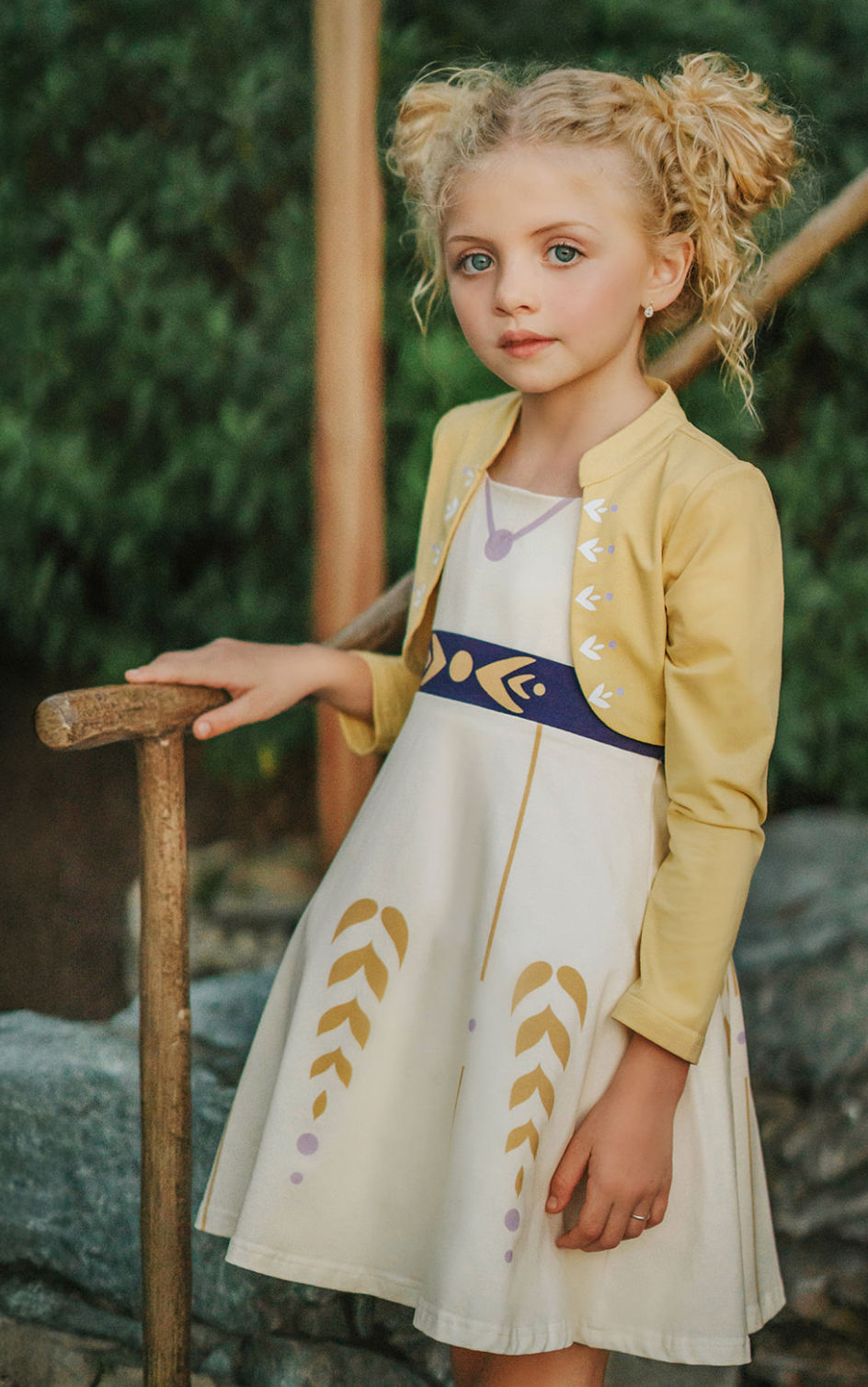 A young girl stands wearing a cotton twirl dress. There is sewn in petti skirt for volume. A geometric golden detail adorns the ivory dress.  There is a purple band with tie at the waist.  The sleevless dress comes with a golden yellow bolero with print on the front in true Arendale princess fashion.