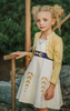 A young girl stands wearing a cotton twirl dress. There is sewn in petti skirt for volume. A geometric golden detail adorns the ivory dress.  There is a purple band with tie at the waist.  The sleevless dress comes with a golden yellow bolero with print on the front in true Arendale princess fashion.