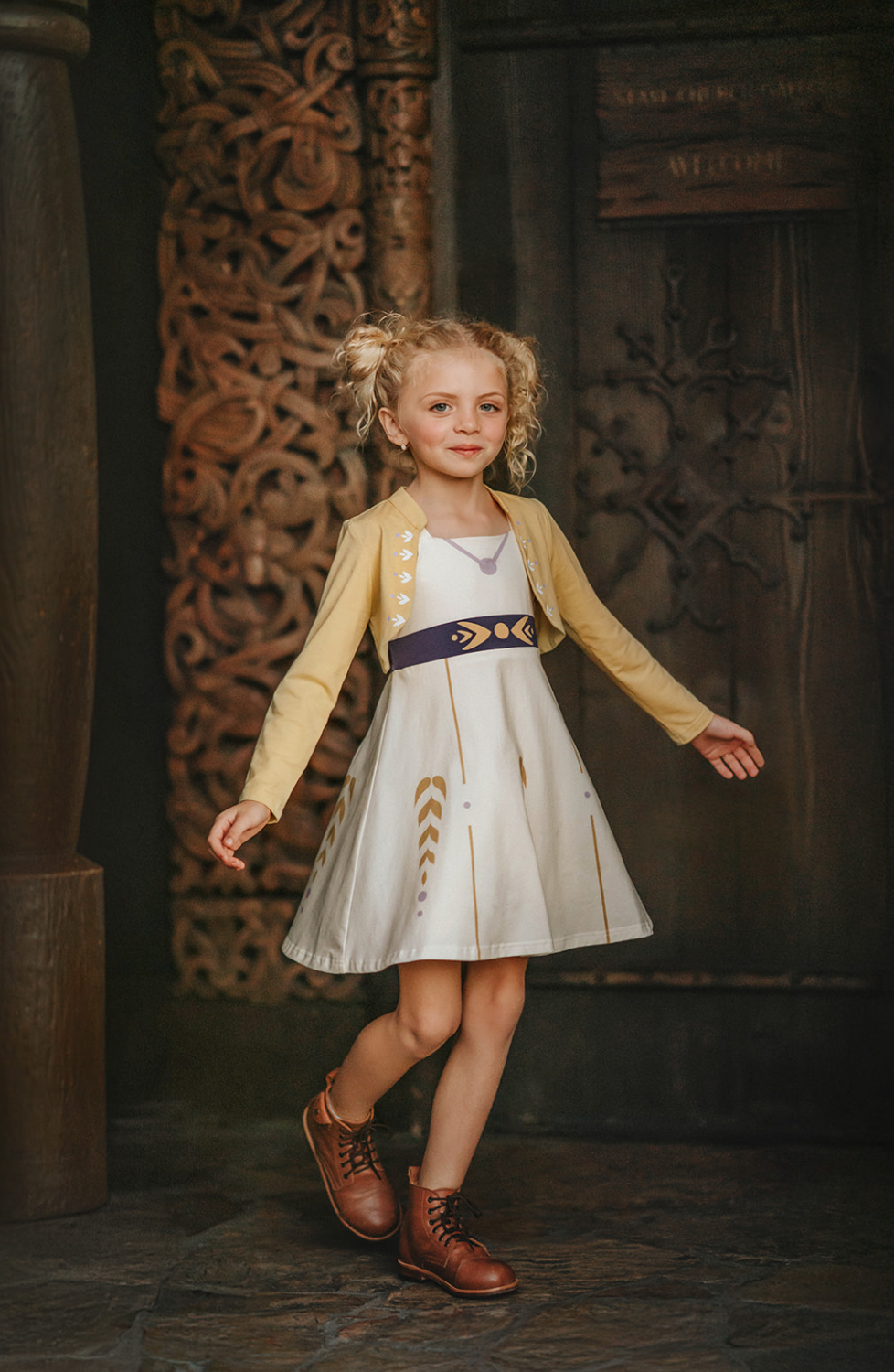 A young girl stands wearing a cotton twirl dress. There is sewn in petti skirt for volume. A geometric golden detail adorns the ivory dress.  There is a purple band with tie at the waist.  The sleevless dress comes with a golden yellow bolero with print on the front in true Arendale princess fashion.