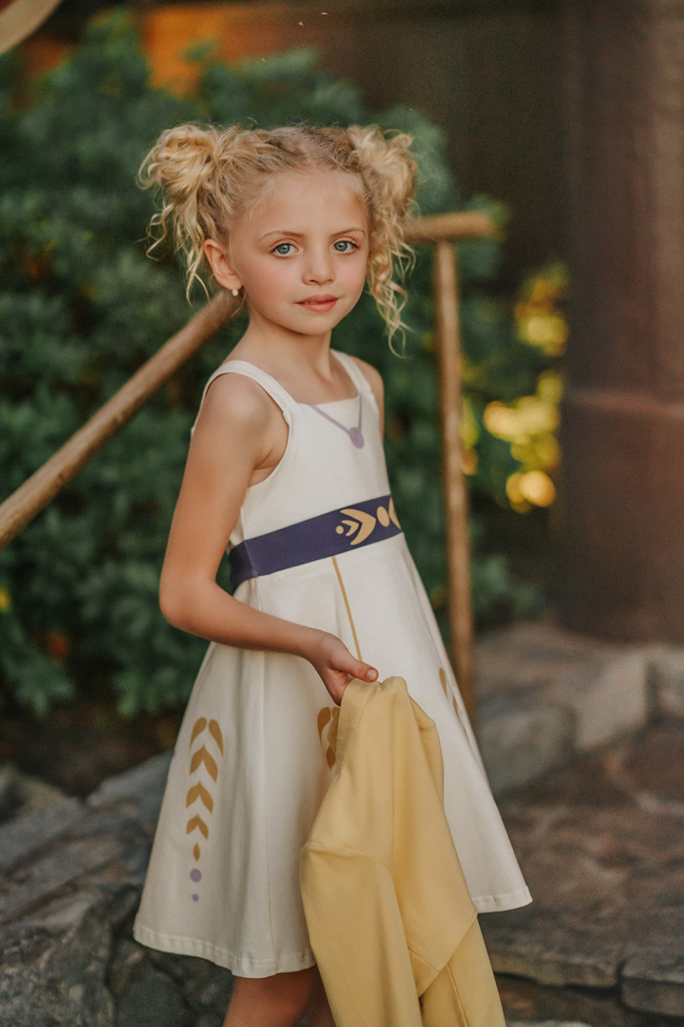 A young girl stands wearing a cotton twirl dress. There is sewn in petti skirt for volume. A geometric golden detail adorns the ivory dress.  There is a purple band with tie at the waist.  The sleevless dress comes with a golden yellow bolero with print on the front in true Arendale princess fashion.