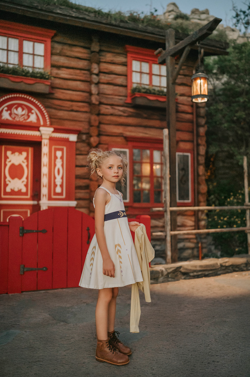 A young girl stands wearing a cotton twirl dress. There is sewn in petti skirt for volume. A geometric golden detail adorns the ivory dress.  There is a purple band with tie at the waist.  The sleevless dress comes with a golden yellow bolero with print on the front in true Arendale princess fashion.