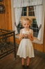 A young girl stands wearing a cotton twirl dress. There is sewn in petti skirt for volume. A geometric golden detail adorns the ivory dress.  There is a purple band with tie at the waist.  The sleevless dress comes with a golden yellow bolero with print on the front in true Arendale princess fashion.