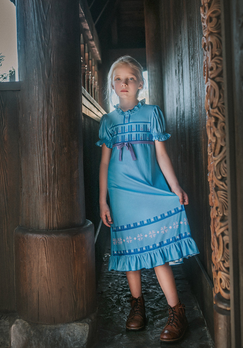 A girl stands in our buttery soft Ice Queen Gown.  A blue nightgown with Scandinavian floral print. Short puff sleeves and a ruffle at the hem.  