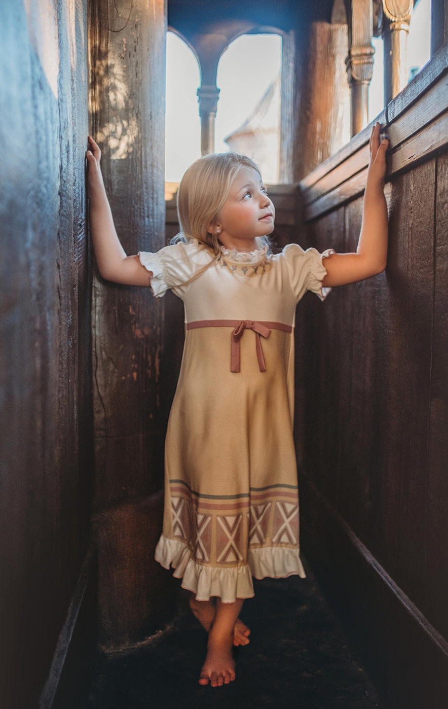 A young girl stands in the little ice princess gown. A light beige and white nightgown with lilac accents. The buttery soft material has geometric prints.  The short puff sleeves are followed are classic with a ruffle at the hem of the gown. 