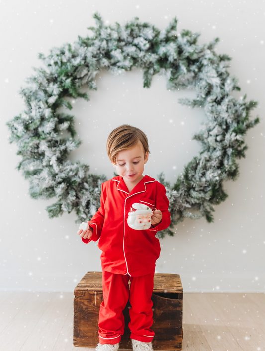 A young child stands on a wooden box, dressed in buttery soft Unisex Pj's in Red from our Christmas Lounge Collection, holding a mug with both hands. Behind them, a large, snowy green wreath hangs on a white wall. Light snowflakes fall gently, adding a festive feel to the scene.