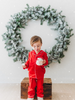 A young child stands on a wooden box, dressed in buttery soft Unisex Pj's in Red from our Christmas Lounge Collection, holding a mug with both hands. Behind them, a large, snowy green wreath hangs on a white wall. Light snowflakes fall gently, adding a festive feel to the scene.