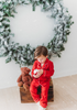 A young child stands on a wooden box, dressed in buttery soft Unisex Pj's in Red from our Christmas Lounge Collection, holding a mug with both hands. Behind them, a large, snowy green wreath hangs on a white wall. Light snowflakes fall gently, adding a festive feel to the scene.