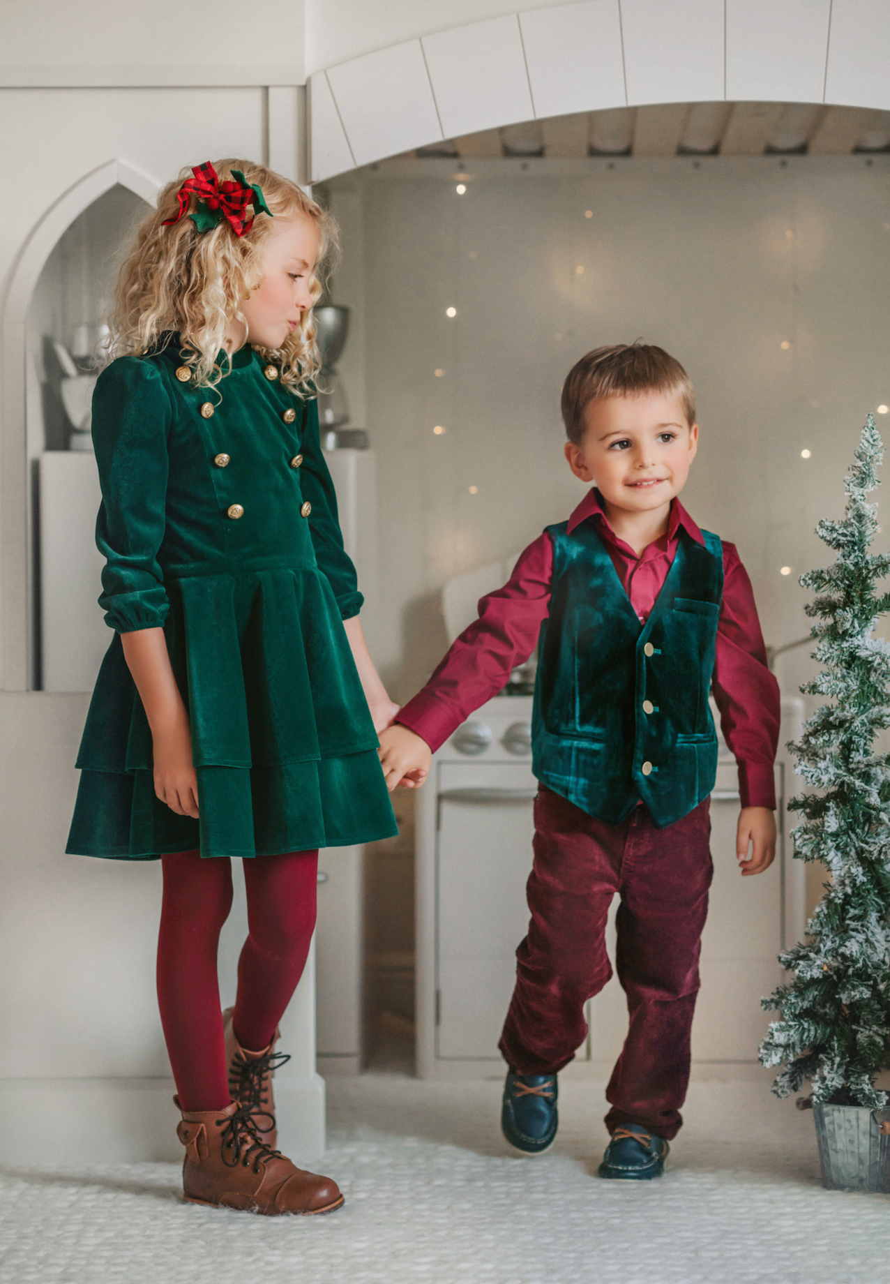 A young boy is with his sibling. He is wearing an evergreen velvet vest with three flat gold buttons.  There is a small pocket on the left chest.  The back has an adjustable buckle to that tightens.  A young girl is wearing the matching evergreen velvet soldier dress. 