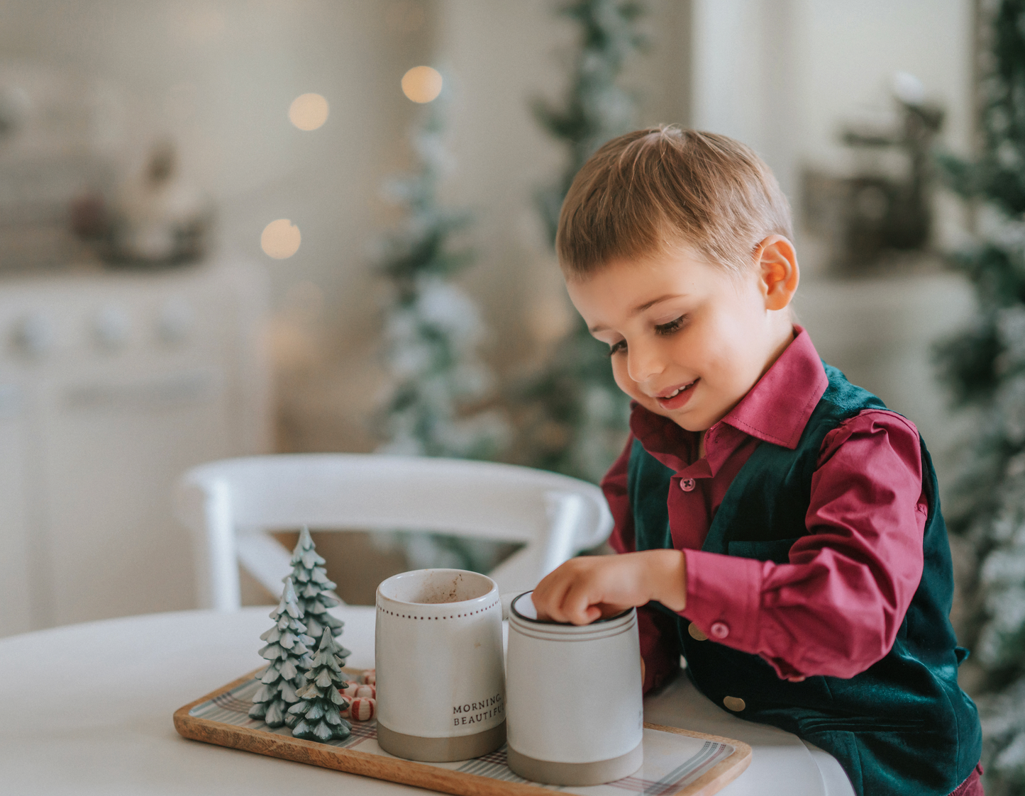 A young boy sits with his hot cocoa. He is wearing an evergreen velvet vest with three flat gold buttons.  There is a small pocket on the left chest.  The back has an adjustable buckle to that tightens.  