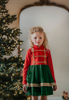 A young girl is wearing a toy soldier twirl dress.  The military collar leads into a cotton bodice adorned with gold thread detail that resembles a soldier uniform.  The evergreen chiffon skirt has multiple layers and is hemmed with gold.  The long sleeve dress is perfect for family holiday photos. 