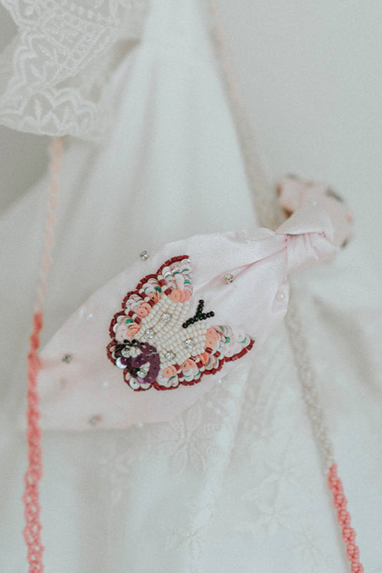 Girl holding a blush pink butterfly headband with beaded detailing, styled in front of a vintage mirror in a lace dress

Close-up of child's hands holding a pink satin headband with intricate butterfly beadwork and pearl accents

Elegant butterfly headband with pastel beads and embroidery, perfect for flower girls, birthdays, or dress-up play