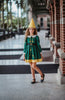 A young girl stands wearing the Smiling Elf Dress, adorned with yellow embroidery, a white collar, and a yellow hem. She also sports a matching yellow cone hat.