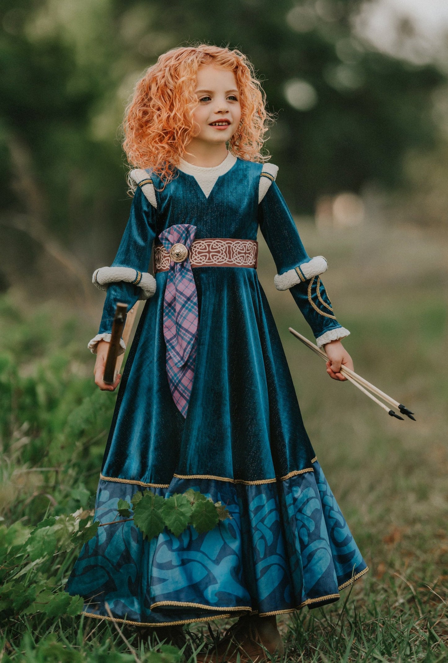 A young girl is wearing a long peacock blue green velvet gown.  The velvet has silver sparkle. The long sleeves have off white shoulder and elbow detail.  There is gold rope trim around the skirt.  A Celtic pattern band in tan has a purple and blue plaid sash with a gold button accent.  The base of the dress has a Celtic inspired pattern in shades of blue green. 