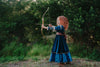 A young girl is wearing a long peacock blue green velvet gown.  The velvet has silver sparkle. The long sleeves have off white shoulder and elbow detail.  There is gold rope trim around the skirt.  A Celtic pattern band in tan has a purple and blue plaid sash with a gold button accent.  The base of the dress has a Celtic inspired pattern in shades of blue green. 