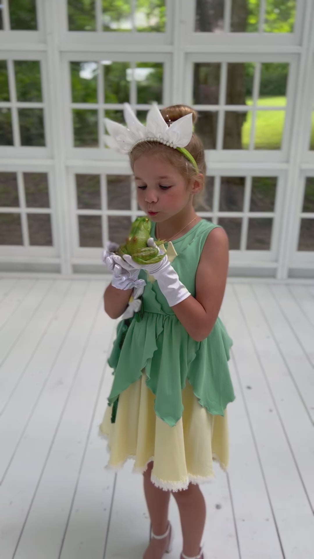 A young girl poses in the Frog Princess Twirl Dress . She wears a green and yellow dress with a magnolia accent at the waist. The light green top and leaf shaped skirt are overlayed on a light yellow skirt with white eyelet trim at the bottom.  The dress is sleeveless and a comfortable cotton. 