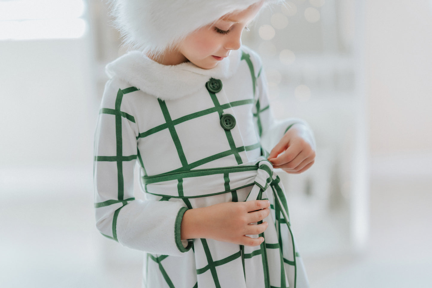 A young girl wears a white and green plaid windowpane dress.  The dress has green buttons at the top and a belt tie.  The collar and cuffs are a soft white fur. 