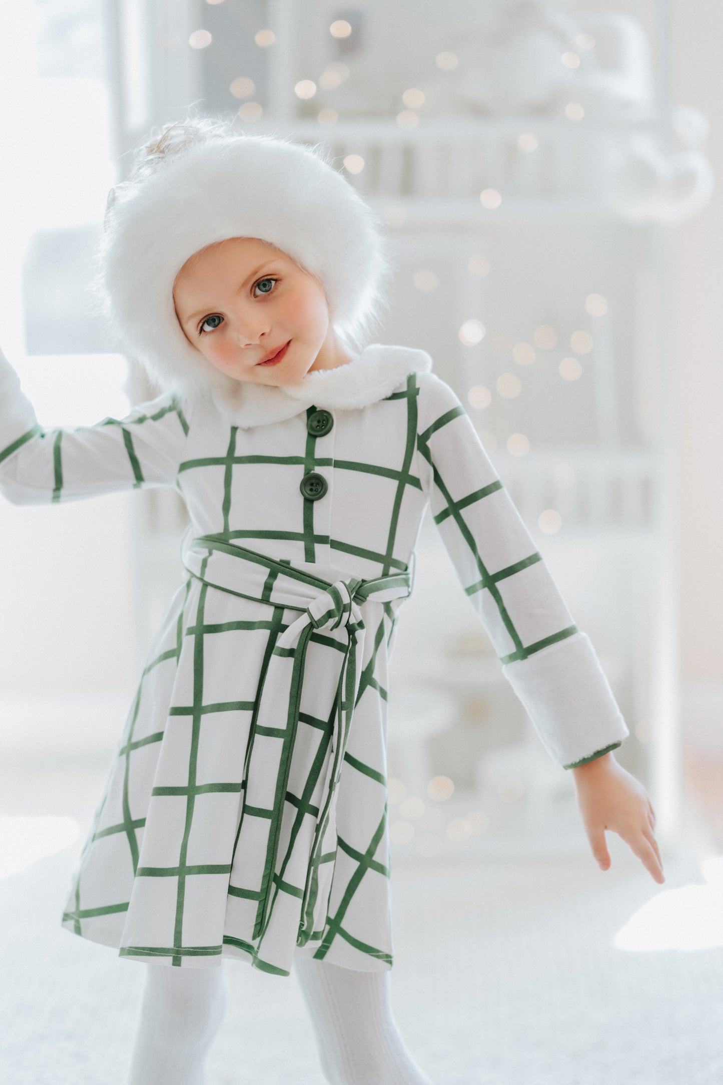 A young girl wears a white and green plaid windowpane dress.  The dress has green buttons at the top and a belt tie.  The collar and cuffs are a soft white fur. 