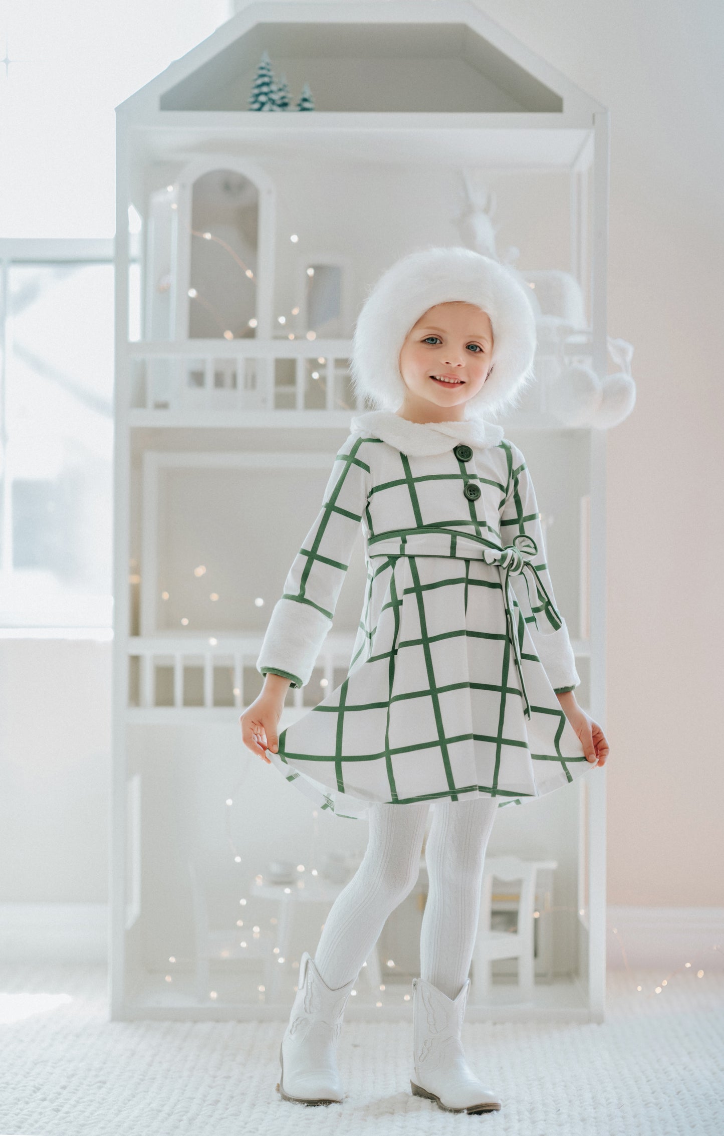 A young girl wears a white and green plaid windowpane dress.  The dress has green buttons at the top and a belt tie.  The collar and cuffs are a soft white fur. 